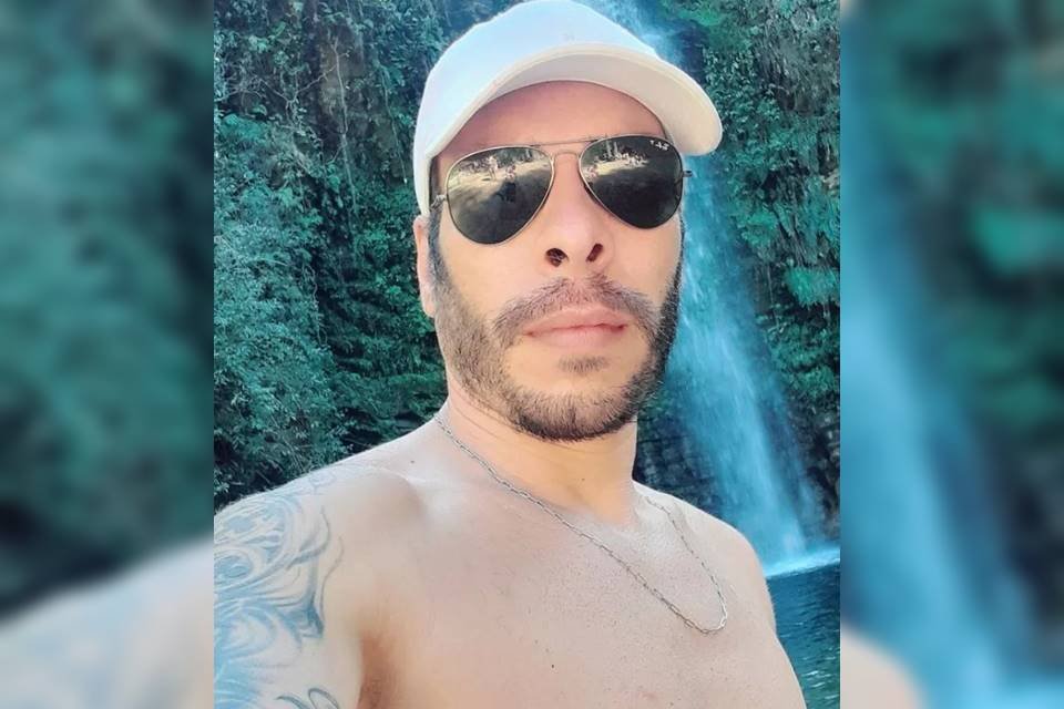 Homem de óculos escuros e boné branco tira selfie em cachoeira - Metrópoles