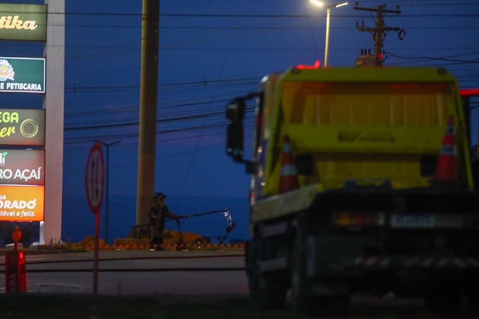 Imagem colorida de área externa, à noite, mostra guincho amarelo, em primeiro plano, com homem ao fundo, vestido com roupa de segurança, enquanto segura, com cabo metálico, item suspeito de ser explosivo