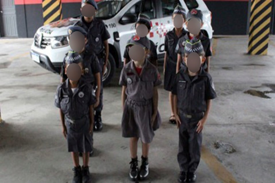 Iamgem colorida mostra oito crianças, com uniformes da PM, enfileiradas na frente de uma viatura - Metrópoles