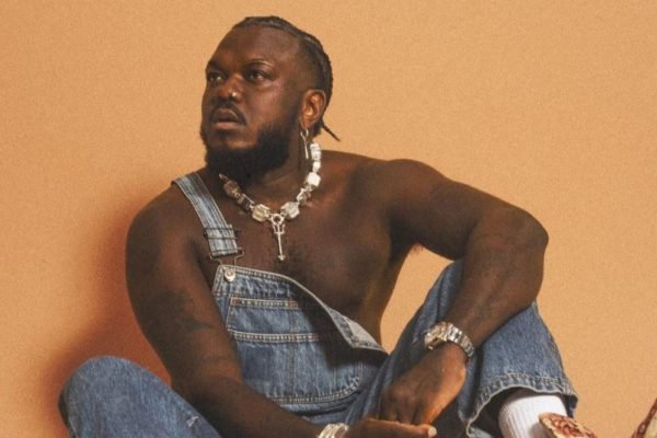 imagem colorida mostra rapper djonga sentado. ele é um homem negro, veste um macacão jeans e usa um colar e um brinco prateados - metrópoles