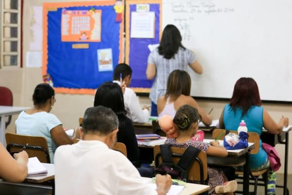 Foto colorida com sala de aula cheia com pessoas maiores de 18 anos e professora escrevendo em lousa