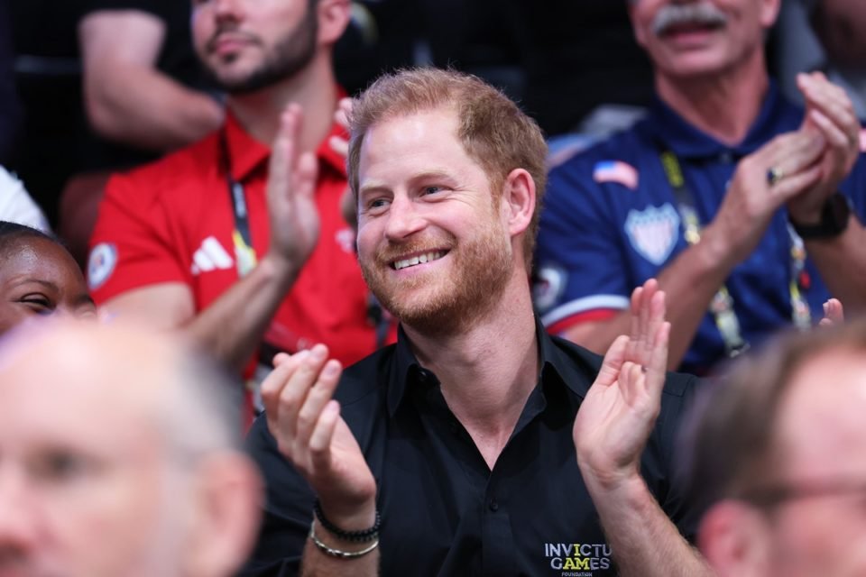 Príncipe Harry, homem branco e ruivo, está sorrindo e aplaudindo algo. Ele está sentando em arquibancada e usando camisa preta - Metrópoles