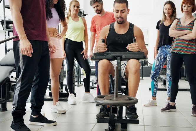 Foto colorida de homem sentado exercitando as panturrilhas com outras pessoas ao redor observando - Metrópoles