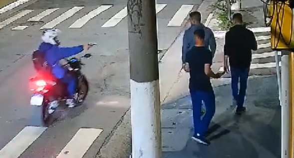 Imagem colorida mostra motoqueiro apontando arma para três homens, que caminham pela calçada - Metrópoles