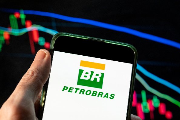 Telefone celular com a imagem do logotipo da Petrobras. Ao fundo, painel com o movimento de ações na Bolsa de Valores - Metrópoles