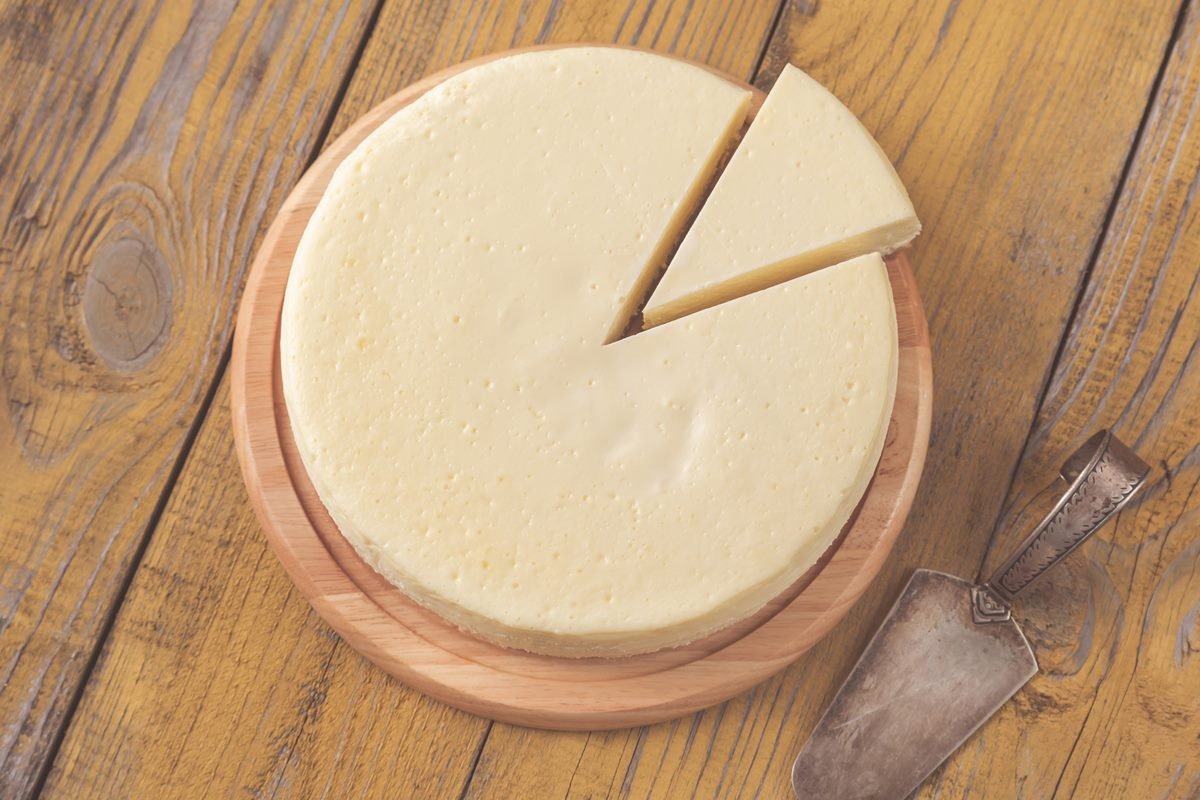 Foto colorida de queijo, com fatia cortada. O alimento está em uma tábua redonda de madeira e perto de uma espátula - Metrópoles