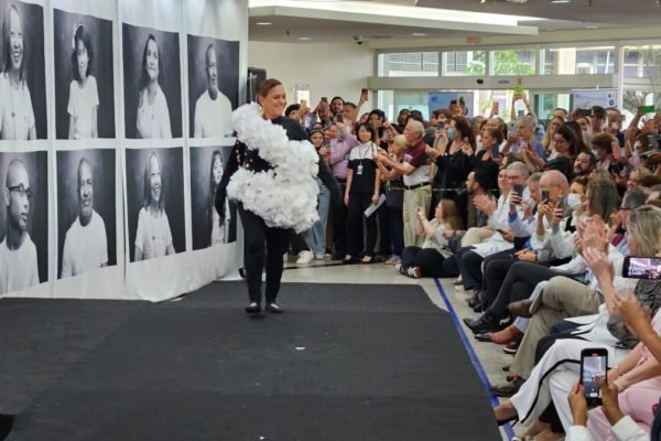 Imagem colorida mostra mulher desfilando em passarela montada em hospital. Ela veste preto e tem uma roupa feita de papel branco por cima do traje. dezenas de pessoas acompanham o desfile e a aplaudem - metrópoles