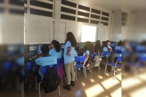 Foto colorida de estudantes com uniforme azul de escola pública olhando para o ocmputador
