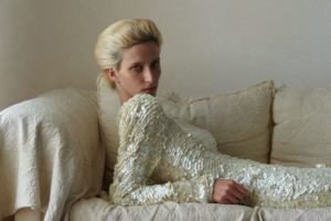 De cabelos loiros, modelo está deitada em sofá com os braços apoiados, usando vestido creme com lantejoulas - Metrópoles
