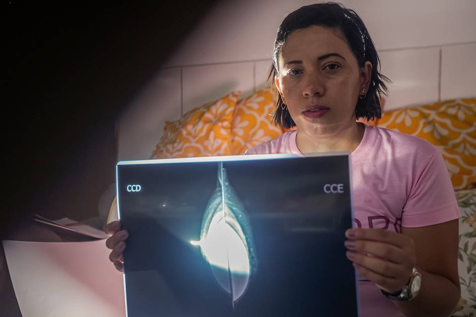 Foto colorida mulher branca com camisa rosa segurando exame com o câncer