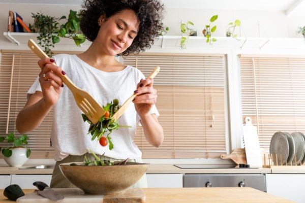 Mulher jovem preparando salada em mesa de cozinha - Metrópoles