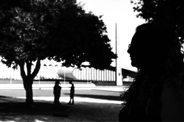 Fotografia em preto e branco mostra perfil de mulher à frente e duas pessoas ao fundo