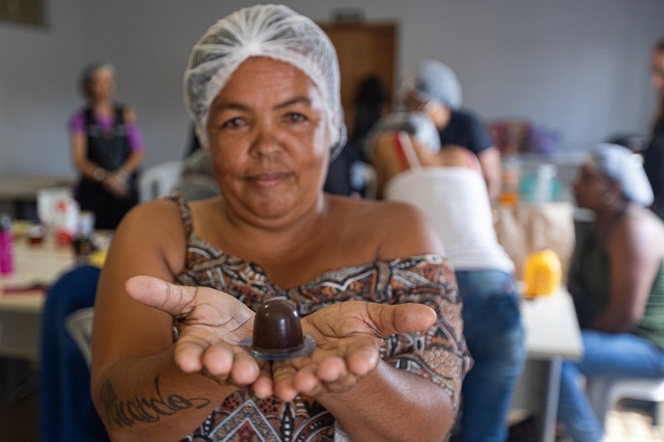 Foto colorida de mulher negra, com chocolate em mãos; ela usa blusa colorida e touca branca no cabelo; ao fundo, há outras mulheres, de costas