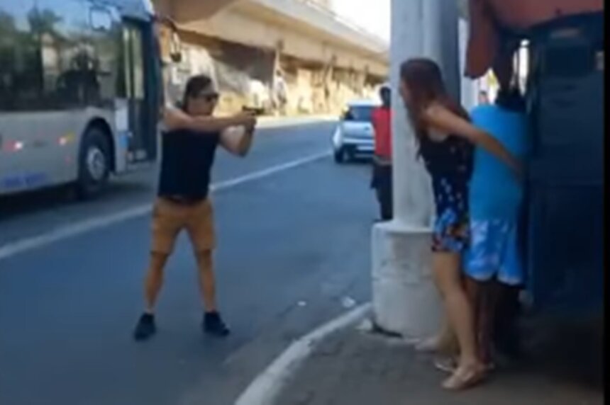 Em foto colorida homem aponta arma de fogo em direção à mulher, que está em frente a jovem - Metrópoles