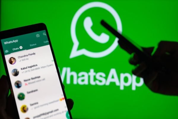 Imagem de uma mão mexendo no celular e outra mostrando um outro aparelho, com mensagens de WhatsApp. Ao fundo, o logotipo do WhatsApp, com as cores verde e branca - Metrópoles