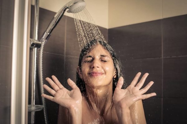 Imagem colorida de mulher mostrando as palmas das mãos, assustada, debaixo de um chuveiro, tomando banho frio - Metrópoles
