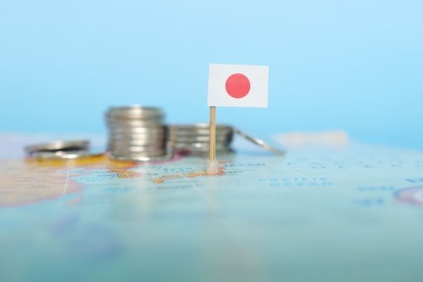 Imagem colorida de uma bandeira do Japão colocada sobre um mapa-múndi, com algumas moedas ao lado, para ilustrar o PIB japonês - Metrópoles