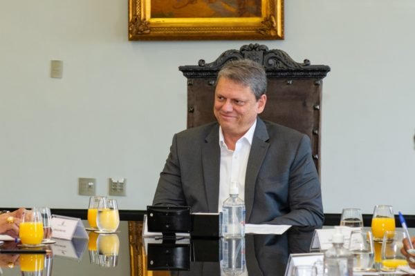 Imagem colorida mostra Tarcísio de Freitas, homem branco, grisalho, vestindo terno cinza e camisa branca, sentado em na ponta de uma mesa de reunião com sucos, papeis e garrafas de água, em uma cadeira de madeira com encosto alto - Metrópoles