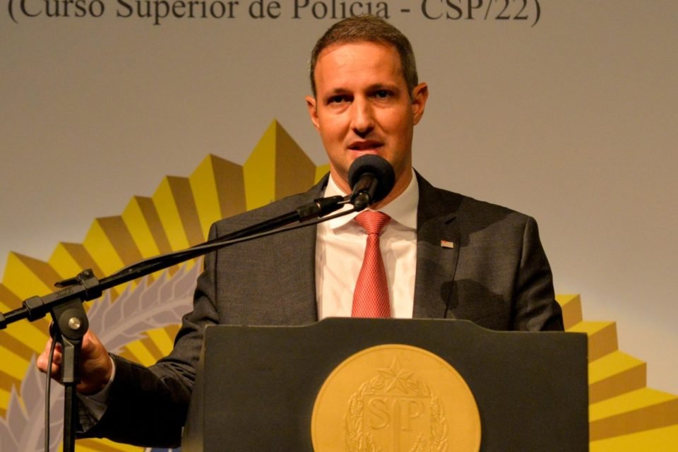 Imagem colorida mostra Guilherme Derrite, homem branco, de cabelo ralo e claro, vestindo terno cinza, camisa branca e gravata avermelhada, falando no microfone em um púlpito do governo de SP - Metrópoles