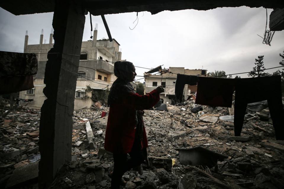 Imagem colorida mostra uma mulher recolhendo as roupas da própria casa, que foi destruída por bobmardeio - Metrópoles