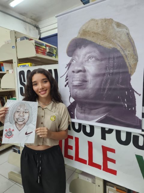 imagem colorida mostra estudante segurando desenho de milton nascimento com camisa do corinthians. ao lado está uma foto do cantor em um banner - metrópoles