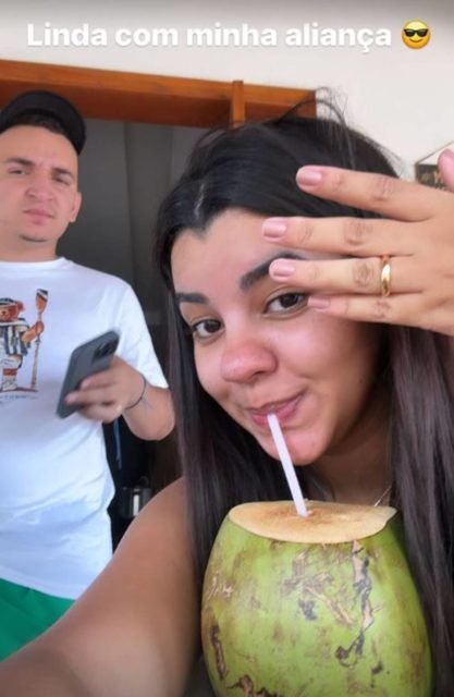 foto colorida de rosto de ary mirelle em primeiro plano tomando agua de coco e com a mão na frente do rosto mostrando uma aliança e joão gomes ao fundo fazendo careta - metrópoles
