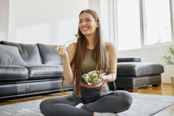 Foto colorida de mulher sentada em um tapete e comendo salada - Metrópoles