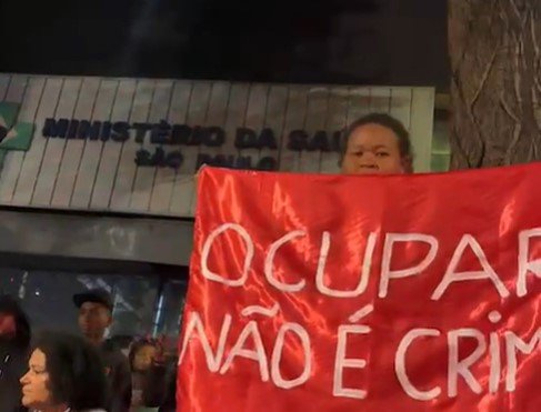 foto colorida de manifestante carregando cartaz em frente a prédio ocupado do Ministério da Saúde em SP - Metrópoles