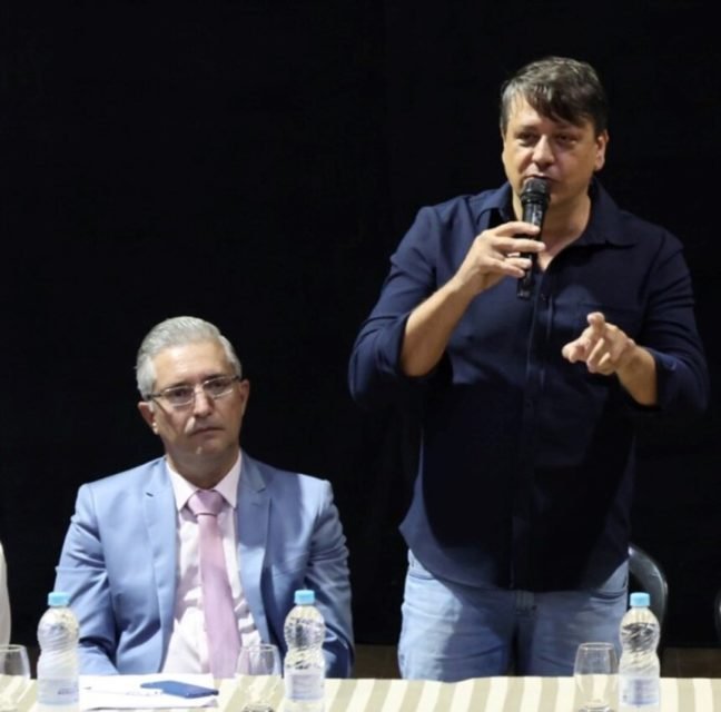 Em foto colorida homem de terno e gravata, sentado, observa outro homem, de camisa e calça jeans, em pé, falando ao microfone - Metrópoles