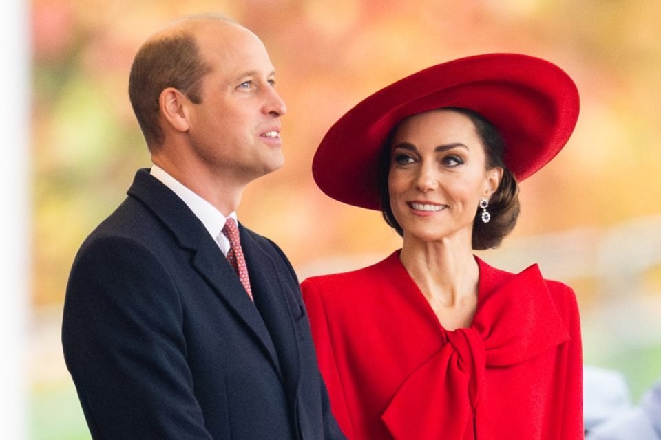 Foto colorida de homem calvo, olhando para cima, vestido com um blazer preto. Ele está ao lado de uma mulher de cabelo castanho preso. Ela usa um chapéu e uma roupa com laço grande na cor vermelha - Metrópoles