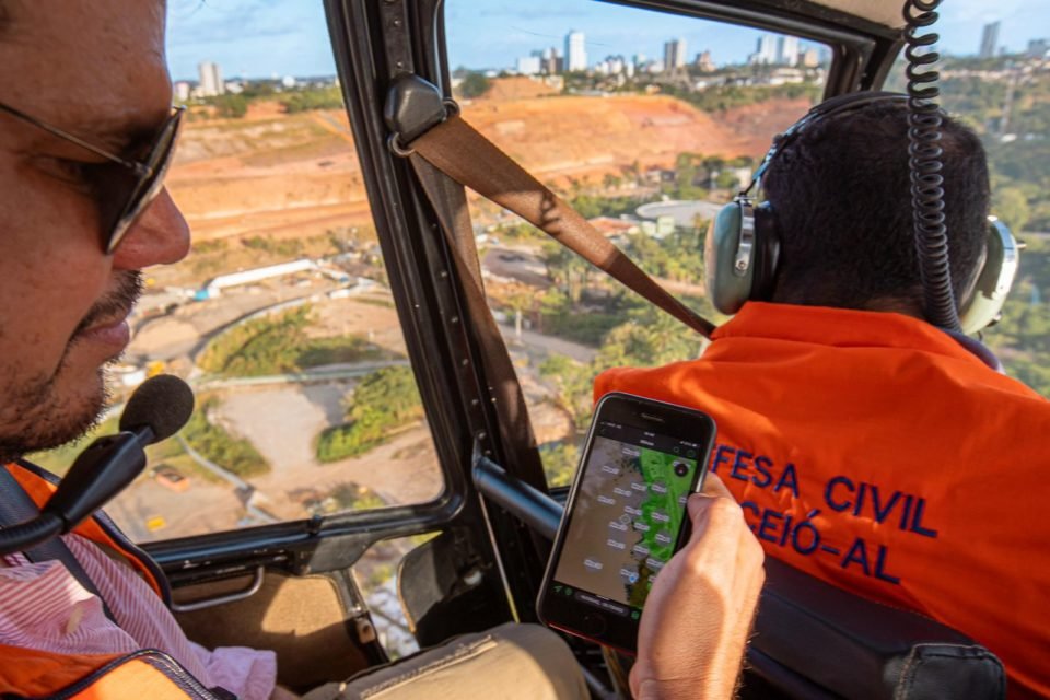 Imagem colorida de homem segurando um celular enquanto sobrevoa local de mina Defesa Civil - Metrópoles