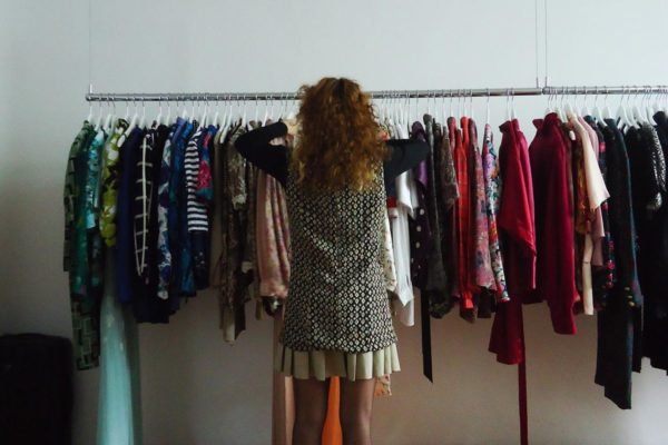 De costas, mulher ruiva de cabelo cacheado mexe em arara de roupas vintage brechó - Metrópoles