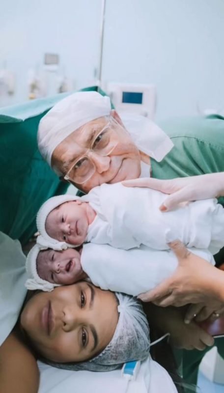 foto colorida de mulher em cama de hospital com duas bebes e um homem com rostos sobre o dela - metrópoles