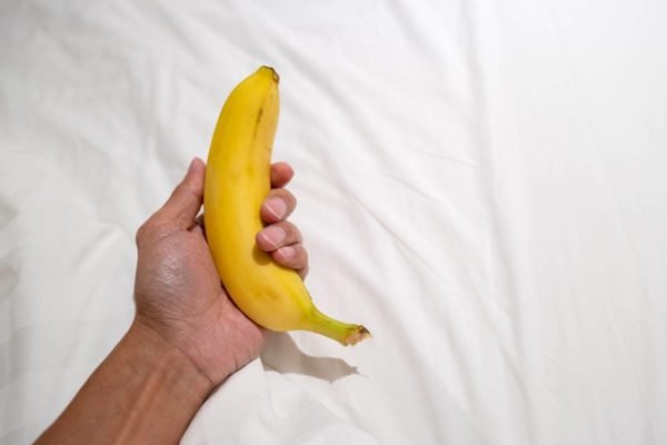 Foto colorida de uma mão masculina segurando uma banana em um fundo branco - Metrópoles