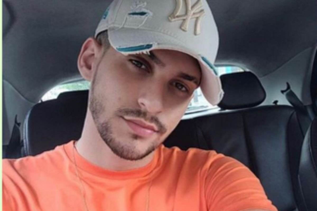 Em foto colorida rapaz usa boné de cor clara, barba rala, e camiseta laranja dentro de carro - Metrópoles
