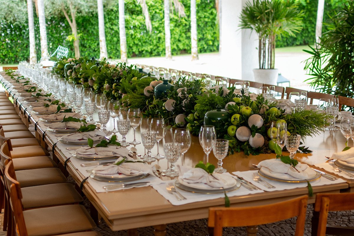 Mesa de almoço com pratos, taças e centro de mesa com folhas e maças verdes - Metrópoles