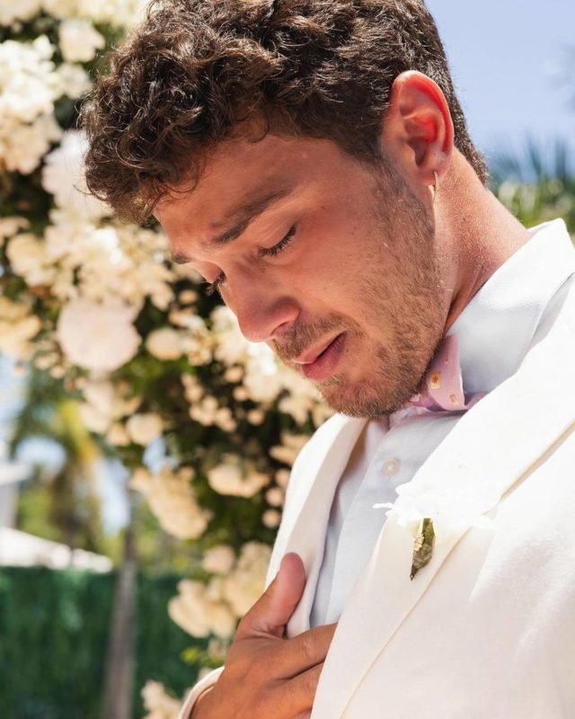 foto colorida de André Luiz Framback de terno branco e gravata borboleta rosa chorando- metrópoles