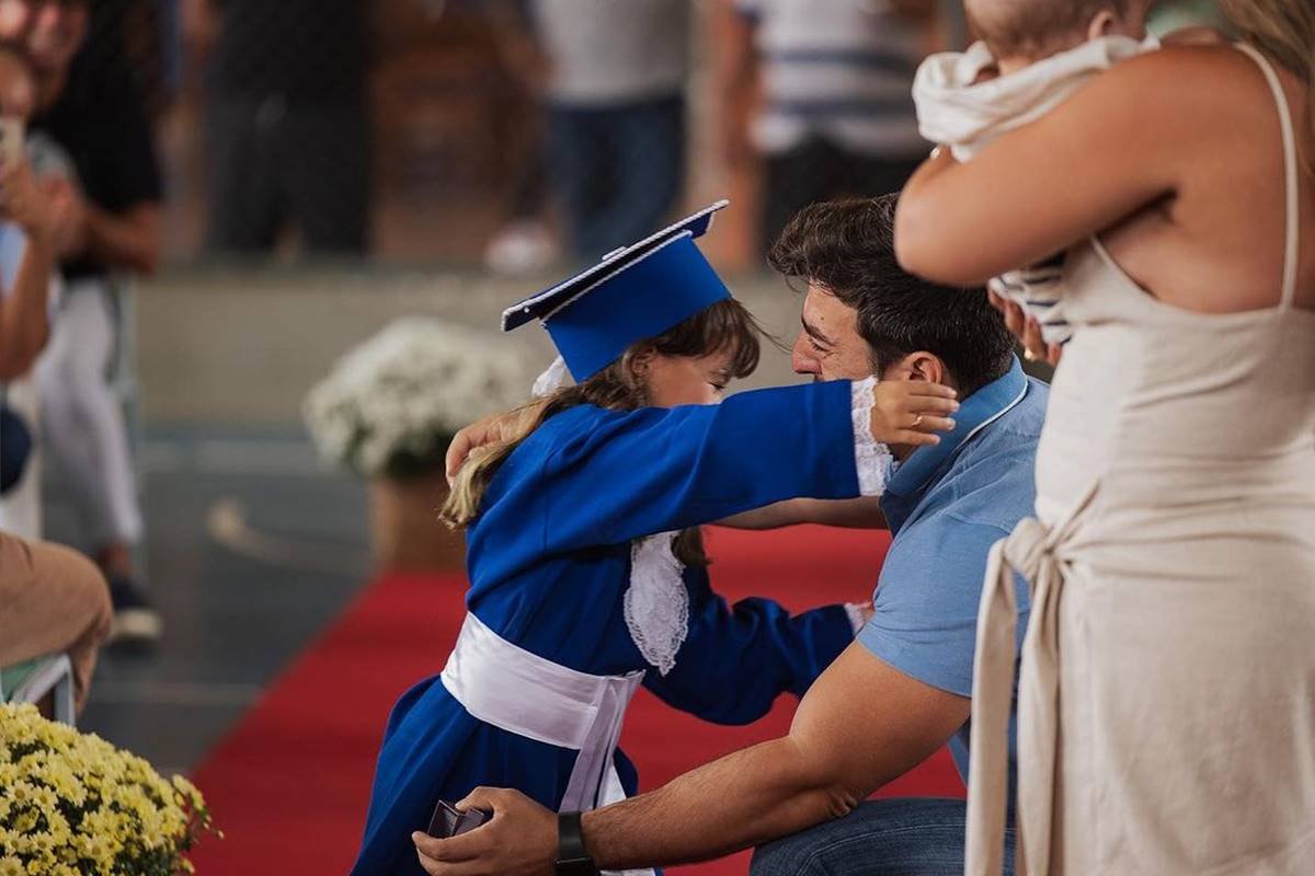Foto colorida de uma criança com roupa de formatura em frente ao pai ajoelhado - Metrópoles