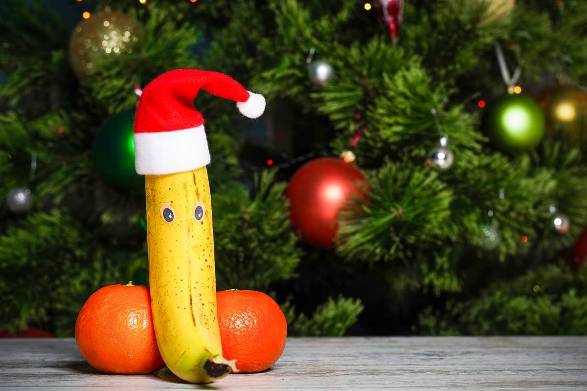 Foto colorida de uma banana com dois olhos e um gorro de papai noel, entre duas tangerinas, em cima de uma mesa e frente a uma árvore de Natal - Metrópoles