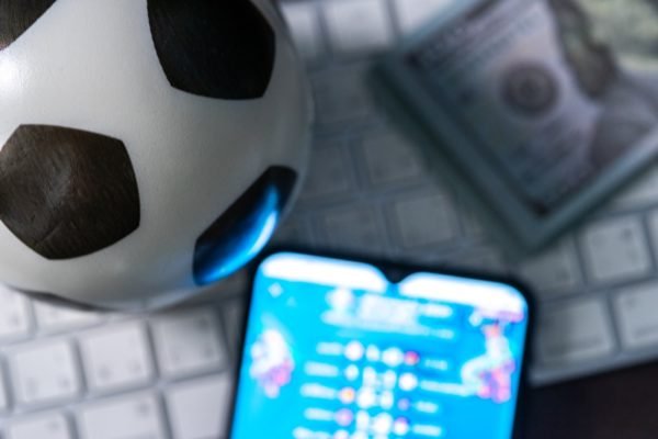 Imagem de telefone celular e bola de futebol sobre um teclado de computador, com notas de dinheiro ao lado - Metrópoles