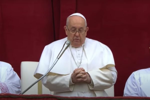 Imagem colorida. Papa Francisco em pé falando em microfone