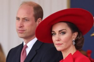 Foto colorida mostra Kate Middleton e príncipe William juntos. Ela, uma mulher branca, com cabelos castanhos, está usando uma vestimenta e chapéu vermelhos. William, homem branco e meio calvo, está usando terno preto e camisa branca - Metrópoles