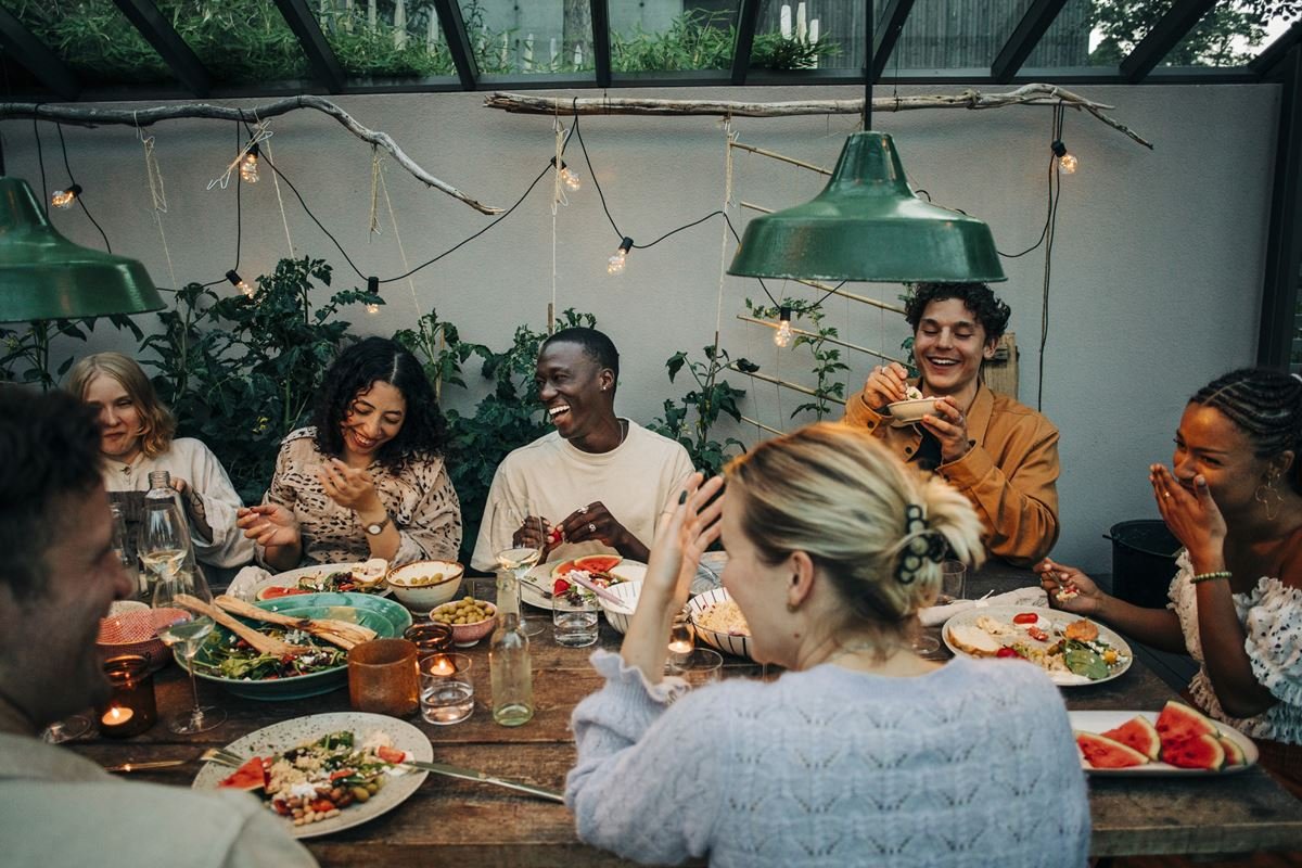 Grupo de pessoas sentado em mesa de refeição. Todos estão sorrindo e interagindo um com o outro - Metrópoles