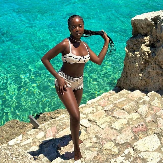 Em escada de pedra, com mar ao fundo, mulher segura as tranças do cabelo longo, usando biquíni com mini shorts de crochê - Metrópoes