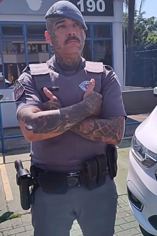 imagem colorida mostra policial militar em frente a base da Polícia. ele veste farda, está com os braços cruzados e tem tatuagens nos braços e rosto - metrópoles