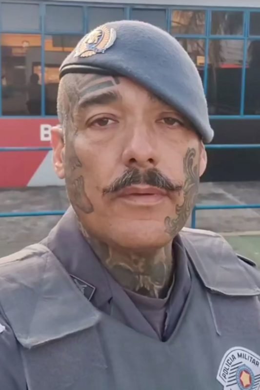 imagem colorida mostra policial militar em frente a base da Polícia. ele veste farda e tem tatuagens no rosto - metrópoles