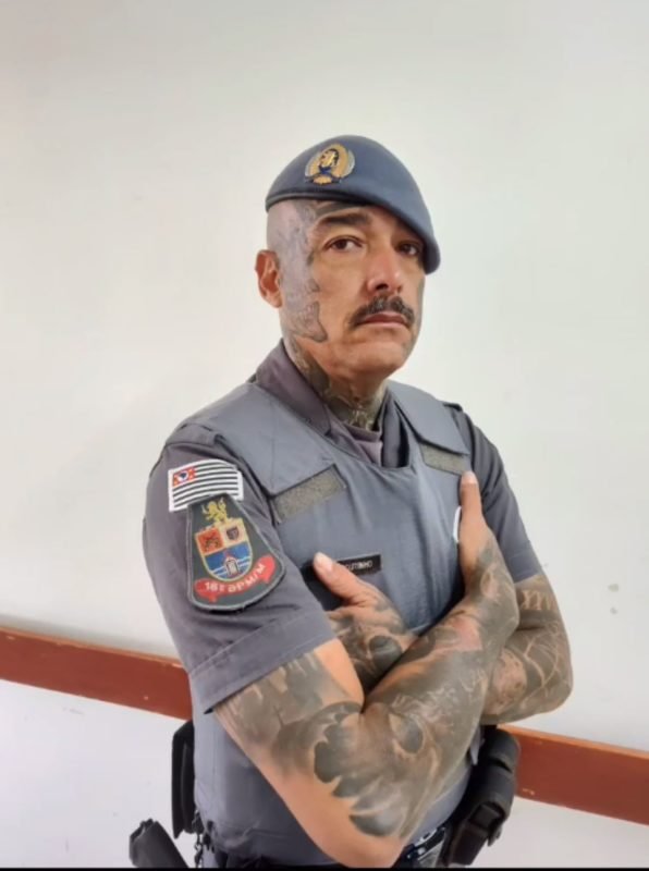 imagem colorida mostra policial militar. ele veste farda, está com os braços cruzados e tem tatuagens nos braços e rosto - metrópoles
