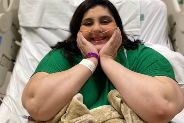 Em foto colorida jovem sorri apoiando ambas as mãos no queixo, usando blusa verde, sobre maca de hospital - Metrópoles