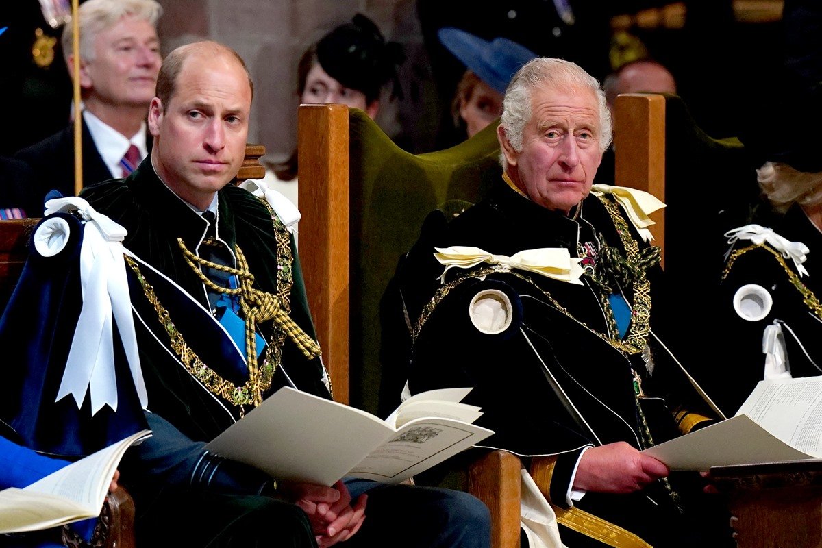 Foto colorida mostra o rei Charles III e príncipe William sentados com roupas formais durante evento. Ambos são brancos e estão olhando sério para o lado - Metrópoles