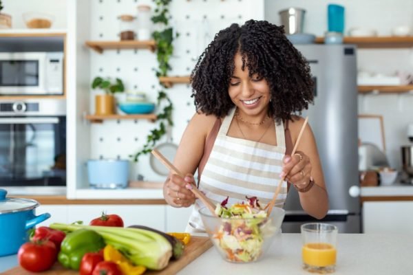 Jovem negra, de cabelos cacheados e sorridente prepara uma saborosa salada na cozinha de casa - Metrópoles